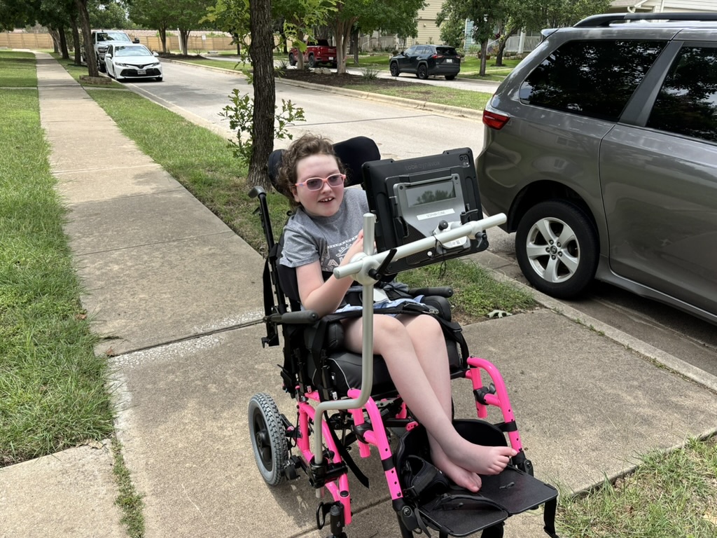 Sophia smiling outdoors in her wheelchair with her communication device